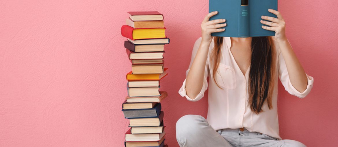 Girl reading a book beside a tall pile of books with a pink wall