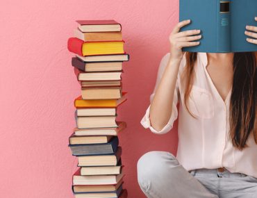 Girl reading a book beside a tall pile of books with a pink wall