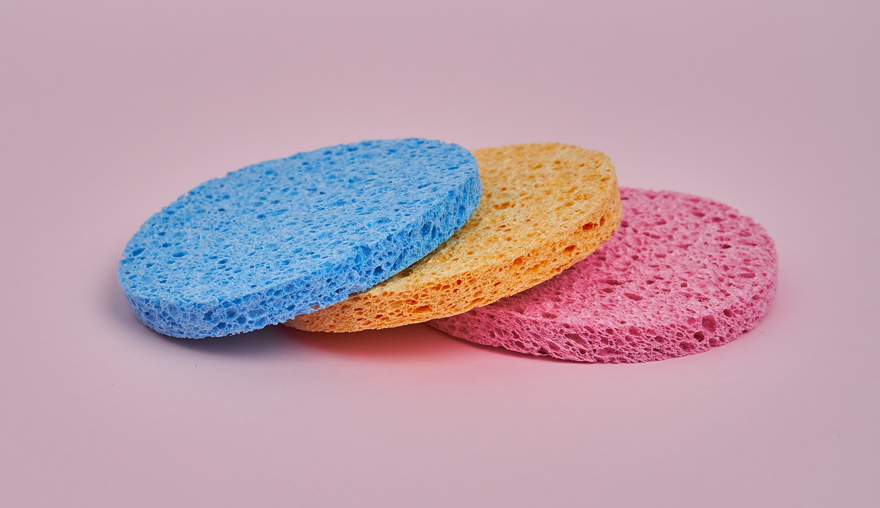 Three flat round sponges (blue, yellow, pink) stacked on top of each other with a pink background