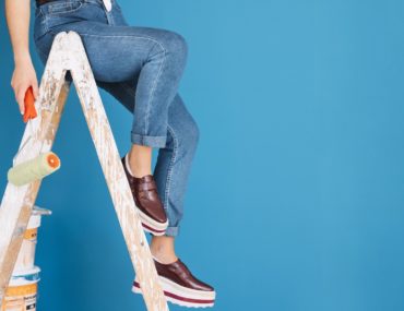 A girl sitting on a ladder with a paint roller in hand with a blue wall behind her