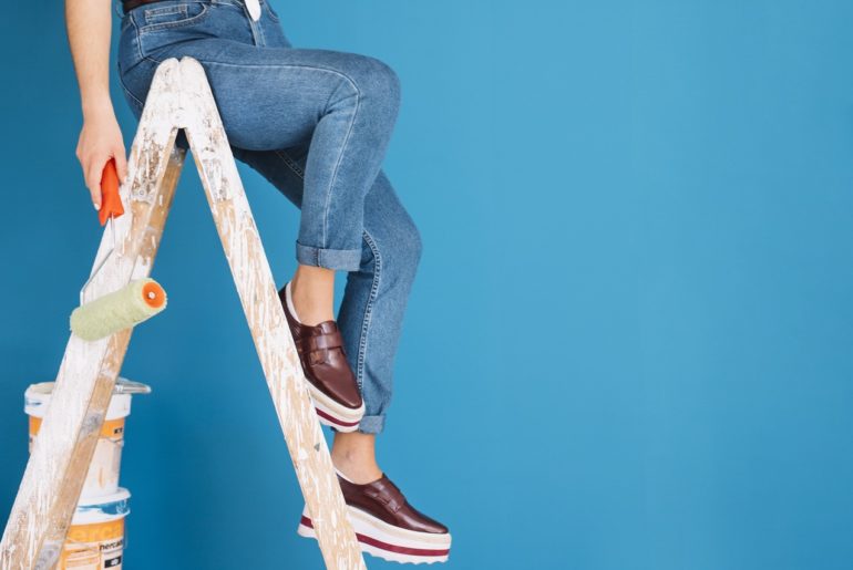 A girl sitting on a ladder with a paint roller in hand with a blue wall behind her